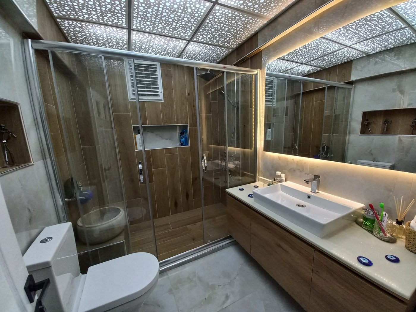 Ahşap ve gri mermer görünümlü fayanslı banyo, aydınlatmalı ayna ve cam duşakabin