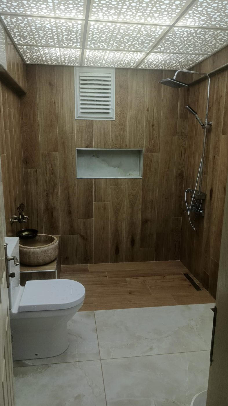 Banyo tadilatı sonrası genel görünüm: ahşap görünümlü fayanslı duş alanı, kurna ve klozet